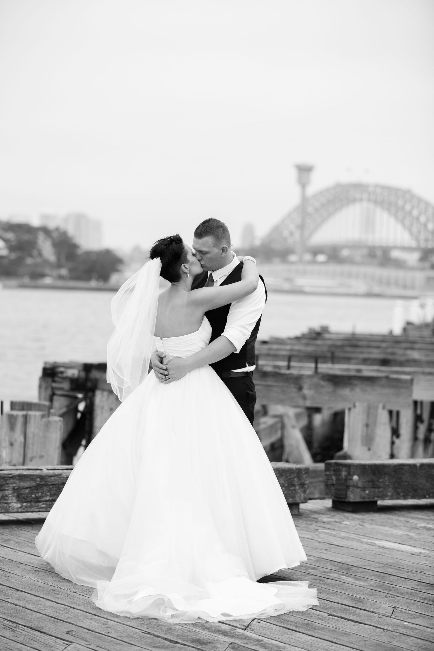Black and white wedding portraits on a waterfront deck with a bridge in the background.