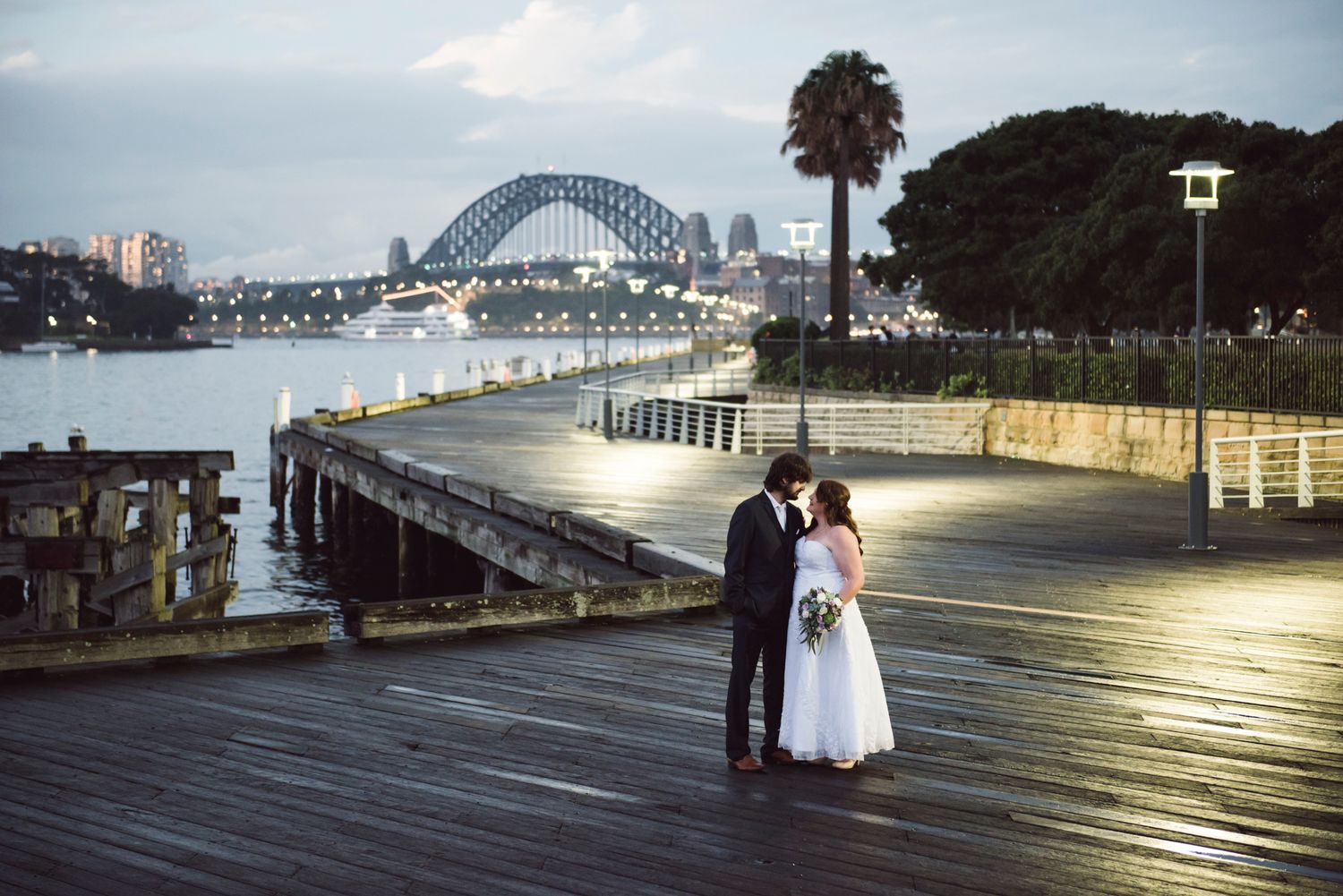 A couple poses on a waterfront boardwalk with Sydney Harbour Bridge in the background at dusk.