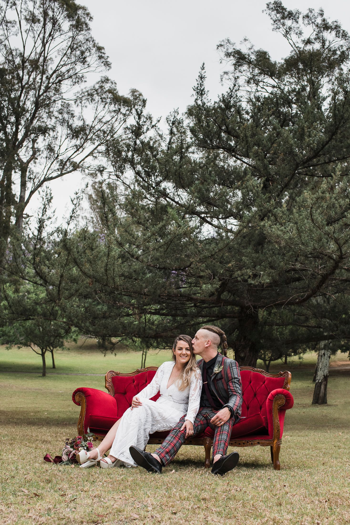A couple sits together on a red vintage couch outdoors surrounded by tall trees in a garden setting.