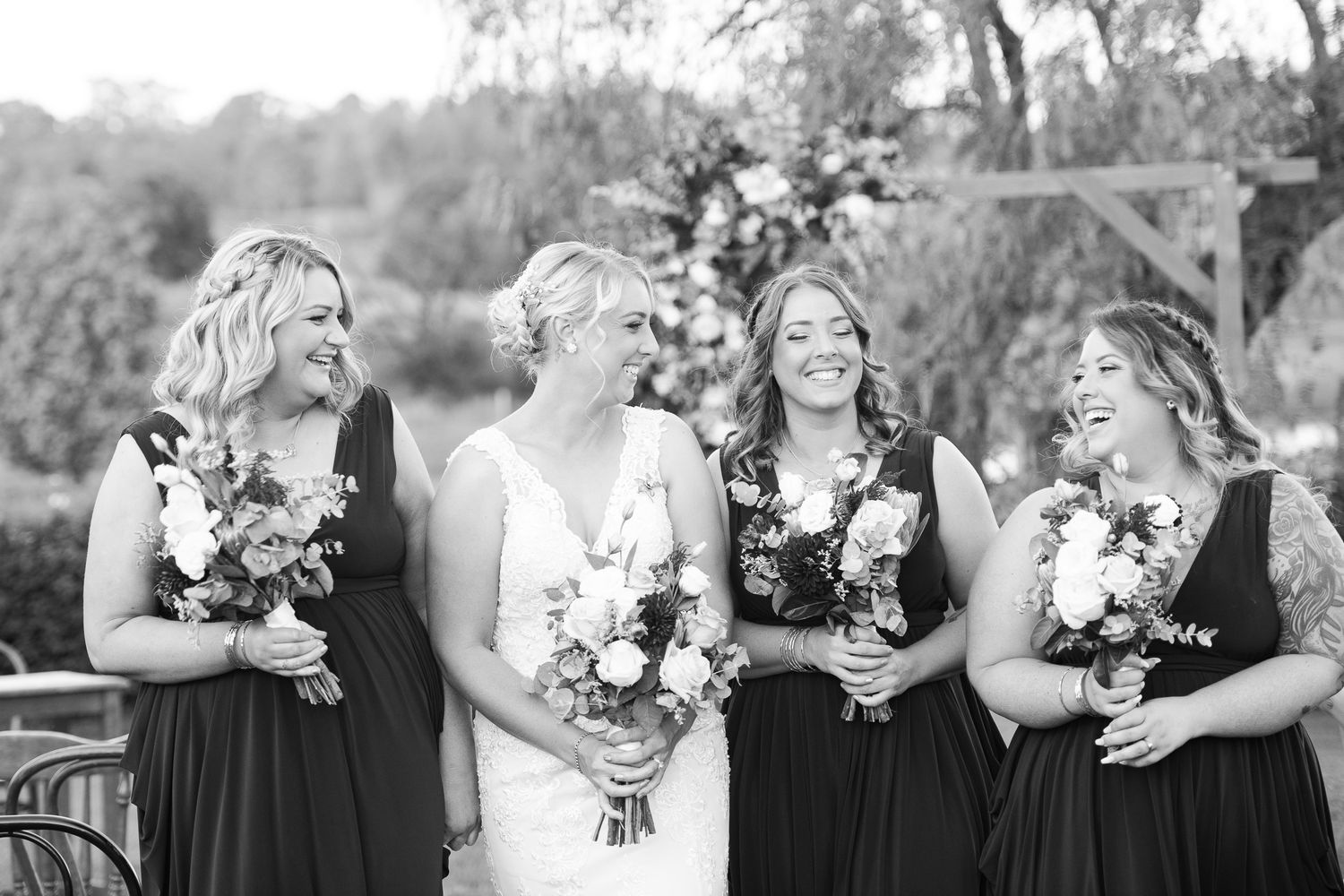 Bride with bridesmaids in black and white formal attire holding bouquets in a garden setting.