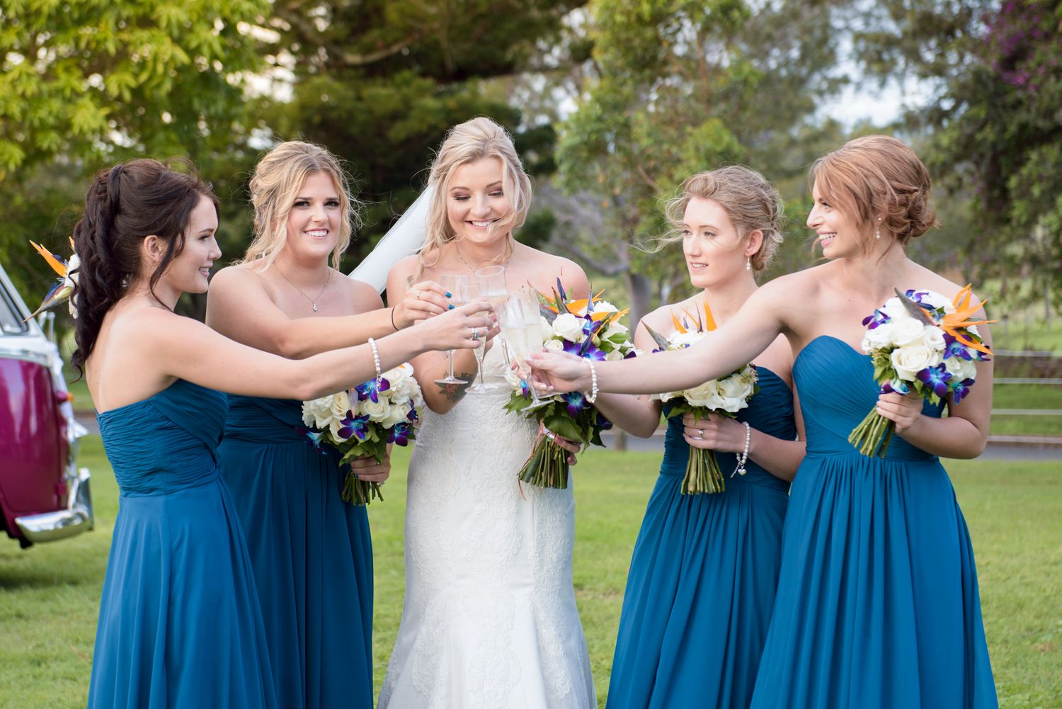 Wedding party in teal blue bridesmaid dresses toasting with bouquets outdoors.