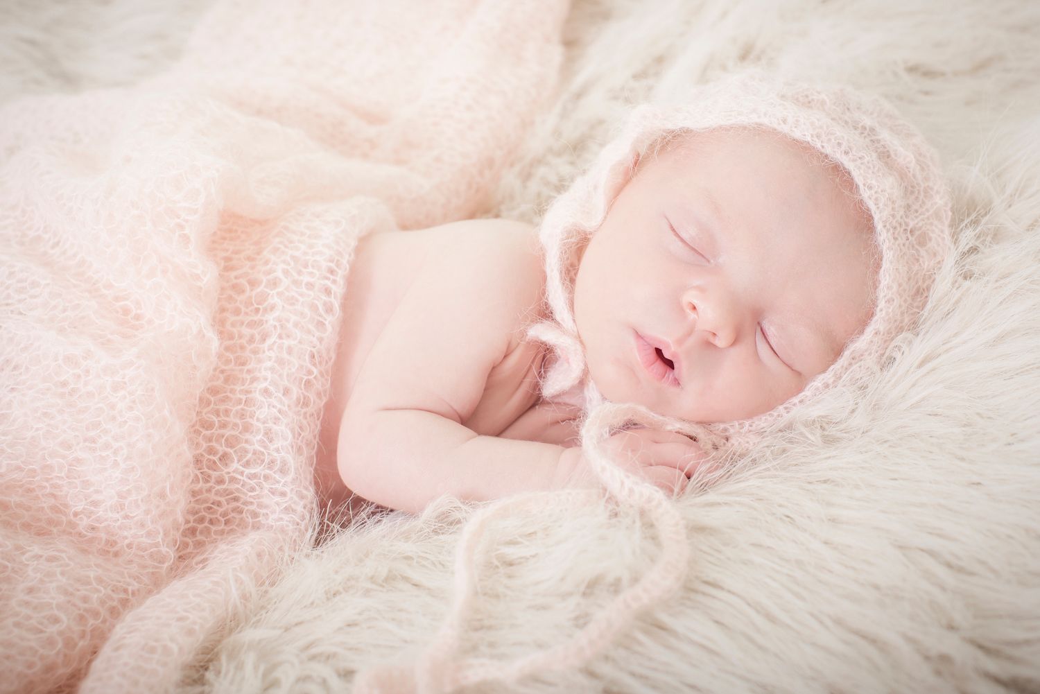 Sleeping newborn on fluffy white fur with soft pink wrap.