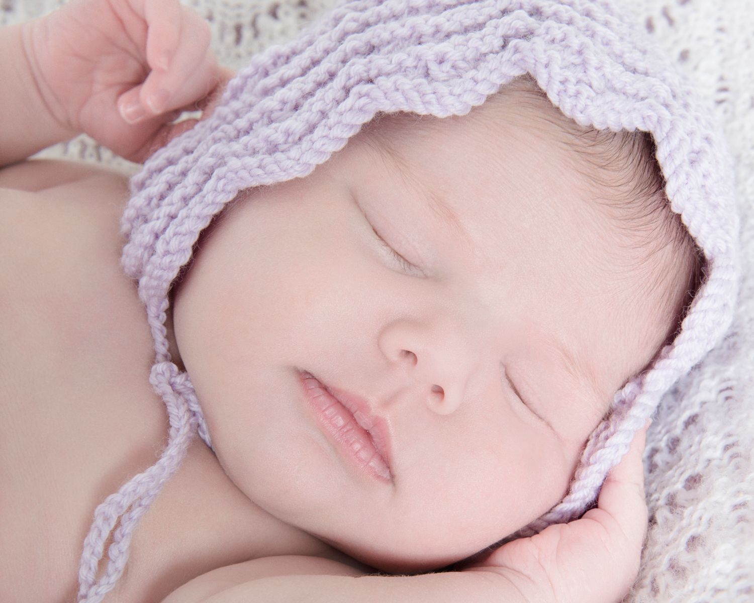 Sleeping newborn with lavender knit cap resting on soft white fabric.