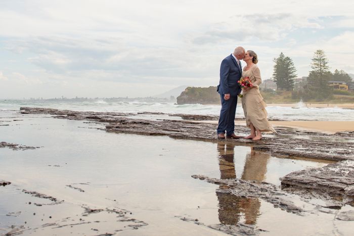 Bride and groom shares a romantic kiss on a rocky beach at sunset with waves crashing in the background.