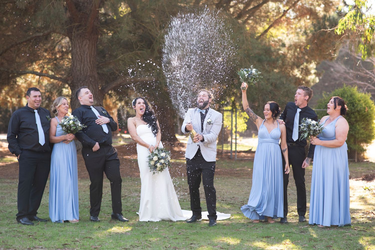 Wedding party in light blue bridesmaid dresses and dark suits poses outdoors in a garden setting.