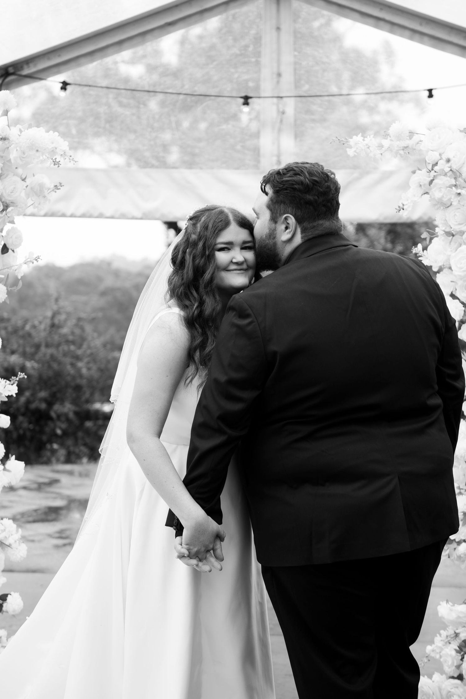 Black and white wedding photo of bride in white gown smiling as groom in black suit whispers in her ear at floral arch.