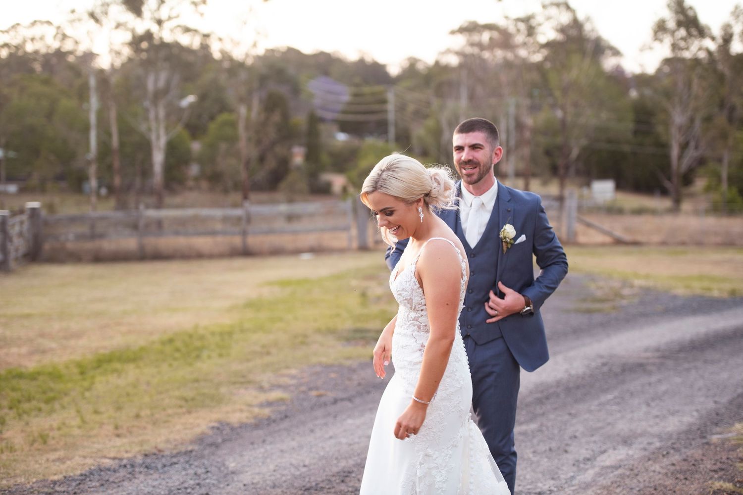 A couple in wedding attire dances and embraces on a rustic dirt path surrounded by trees and natural scenery.