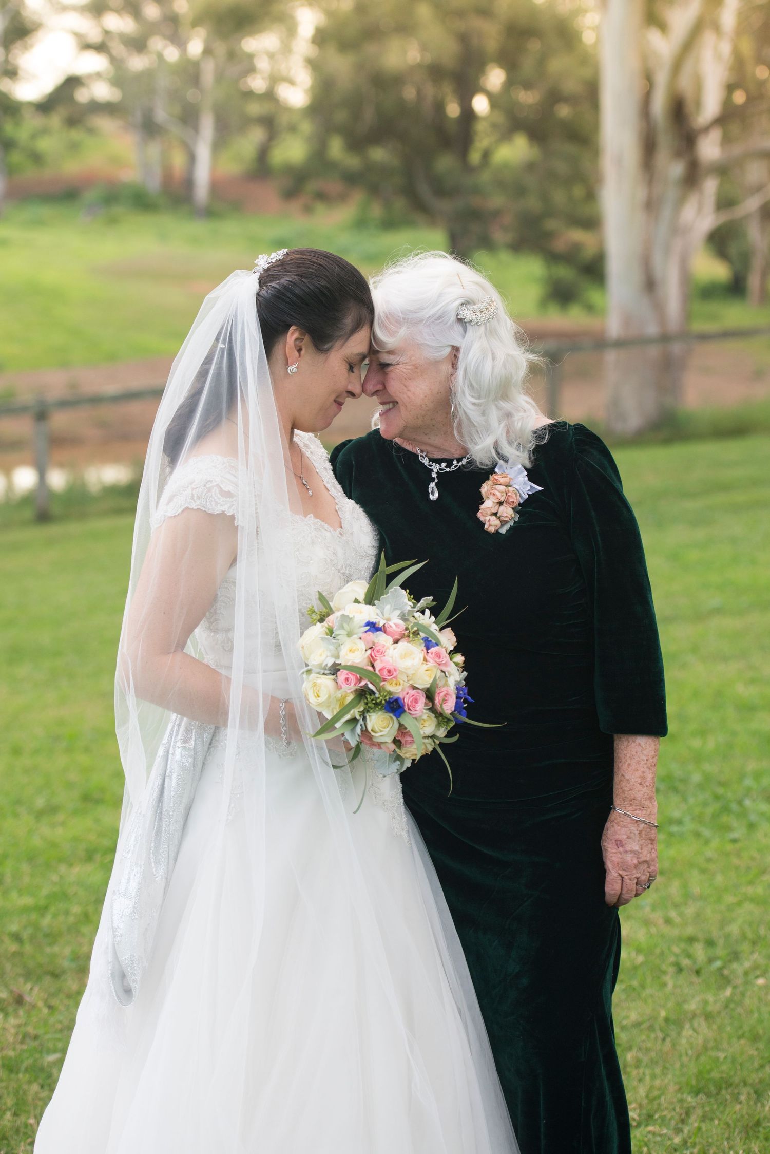 A bride in a white wedding dress shares tender moments with her mother in black during an outdoor ceremony.