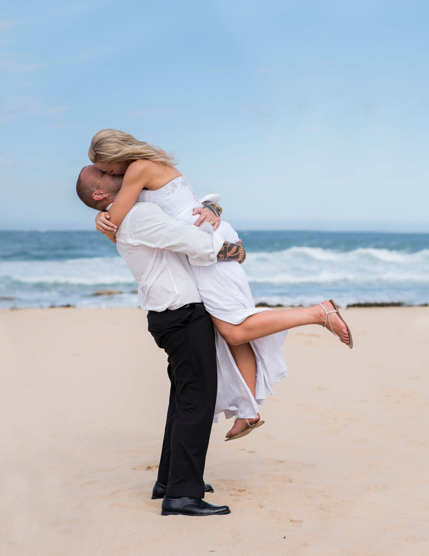 Bride and groom shares a romantic moment on a sunny beach in Wollongong with ocean waves and blue sky in the background.