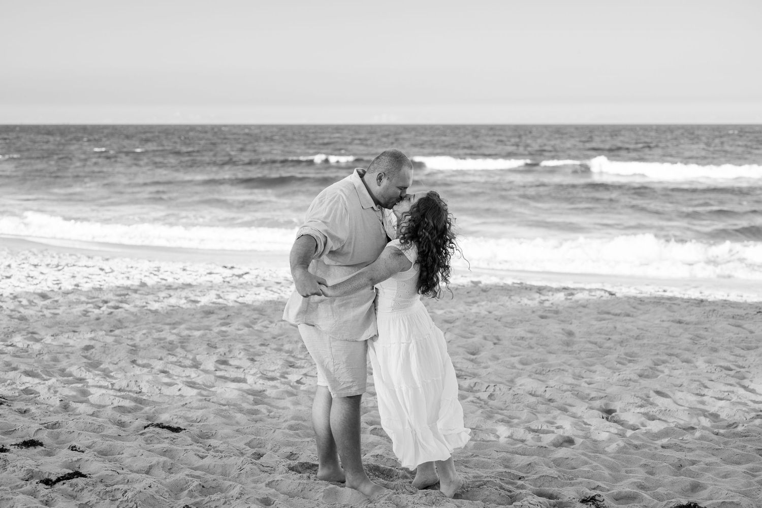 Black and white photo of a couple sharing a romantic kiss on a sandy beach with ocean waves in the background.
