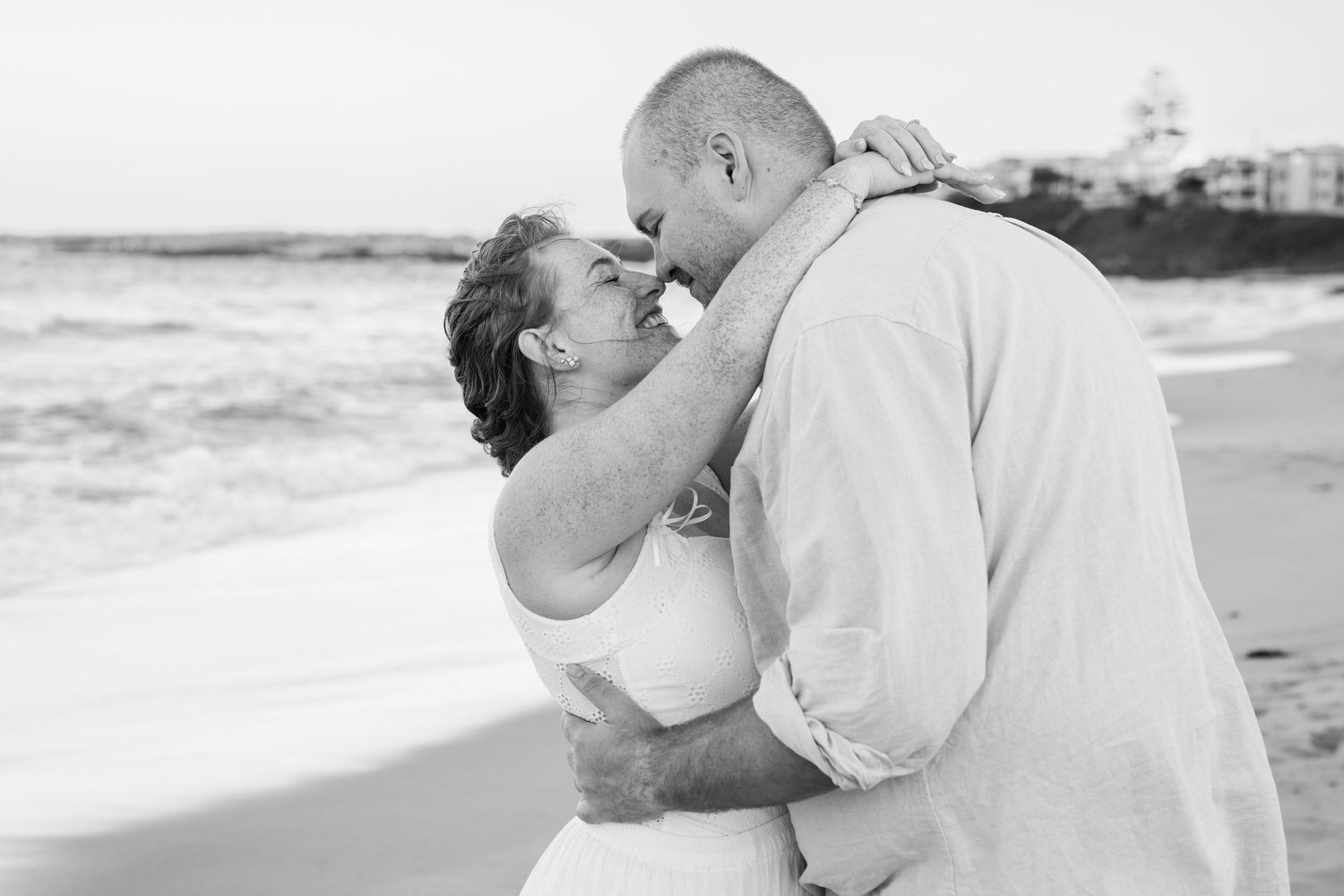 Black and white photo of a couple sharing a romantic embrace on a beach, nearly kissing near ocean waves.