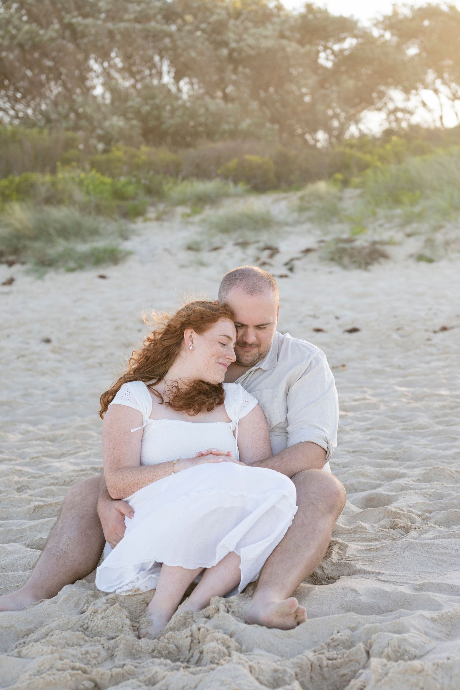 Couple sitting together on sandy beach at sunset, woman in white dress leaning on man, romantic maternity photo.