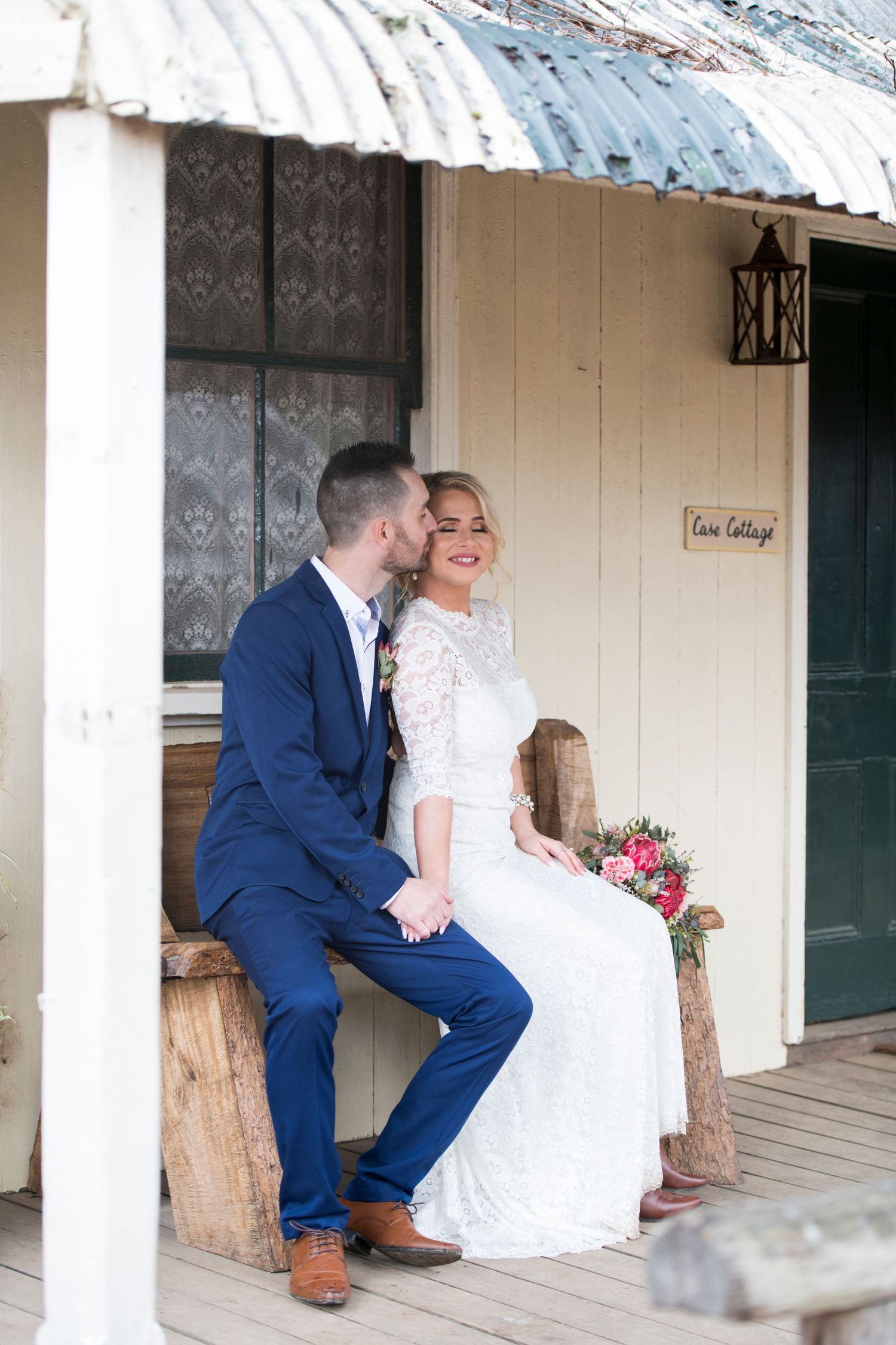 A romantic wedding couple shares intimate moments on a rustic farmhouse porch with white walls and lace curtains.