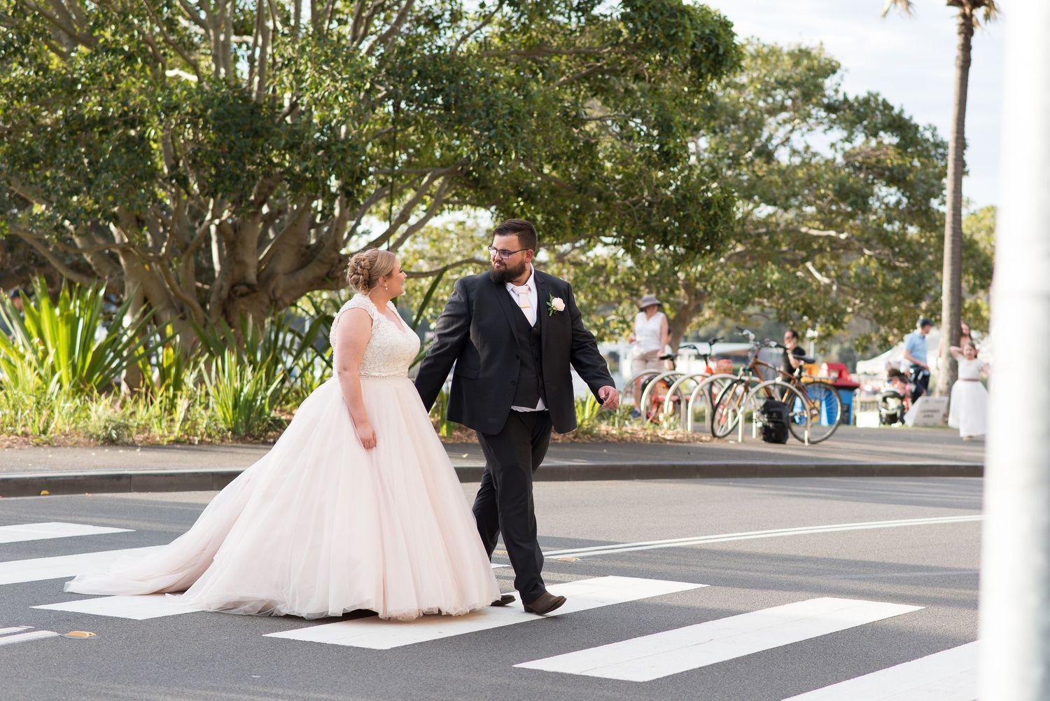 Bride and groom crosses a crosswalk together on a sunny day with trees in the background.