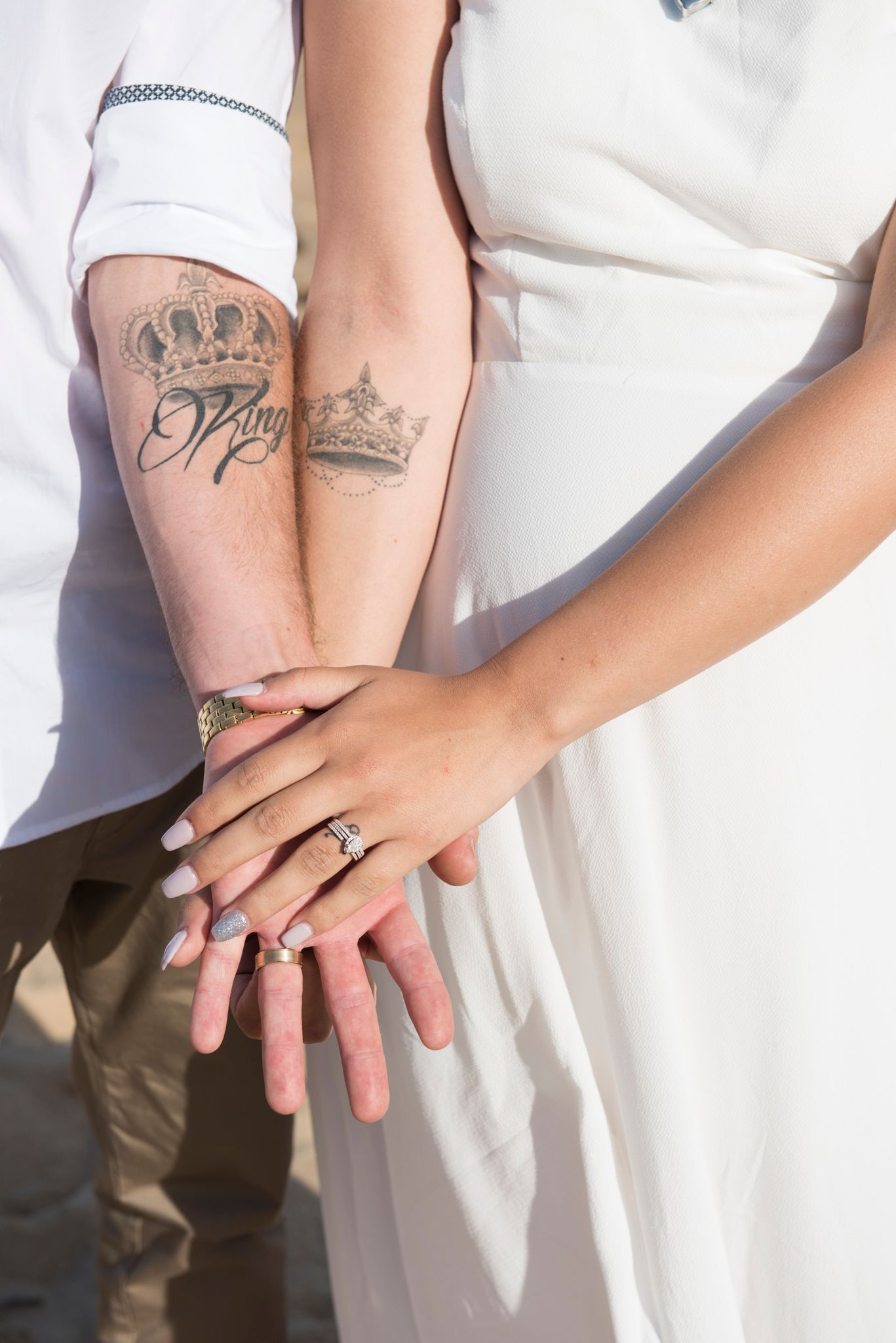 Close-up of couple's hands with tattoos and wedding rings against white dress.