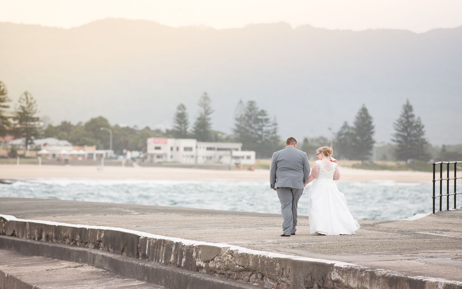 A couple walks along a rocky coastal seawall with waves crashing and beachfront homes visible in the distance.