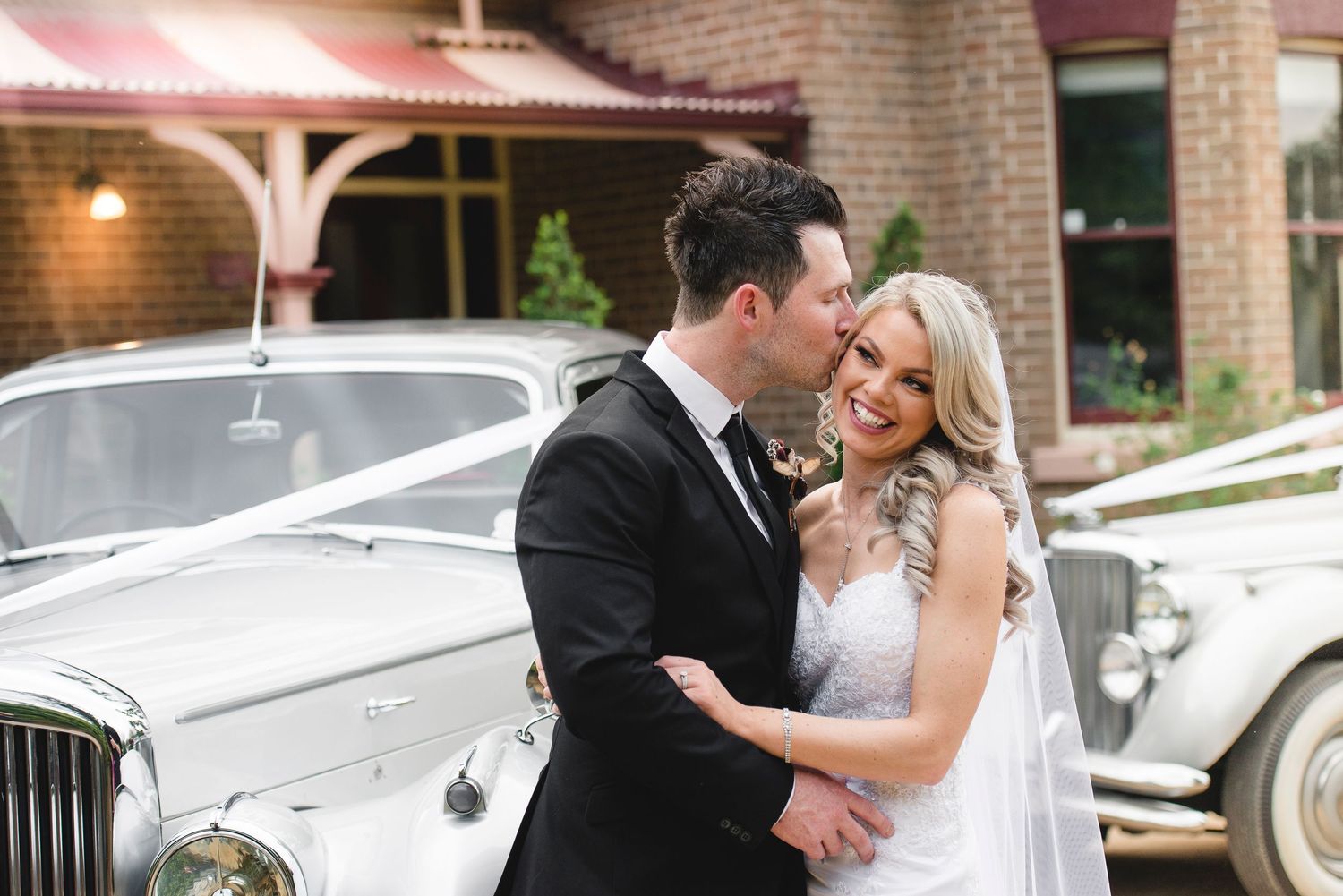 Bride and groom share romantic moments together by white luxury vehicles outside a brick building.