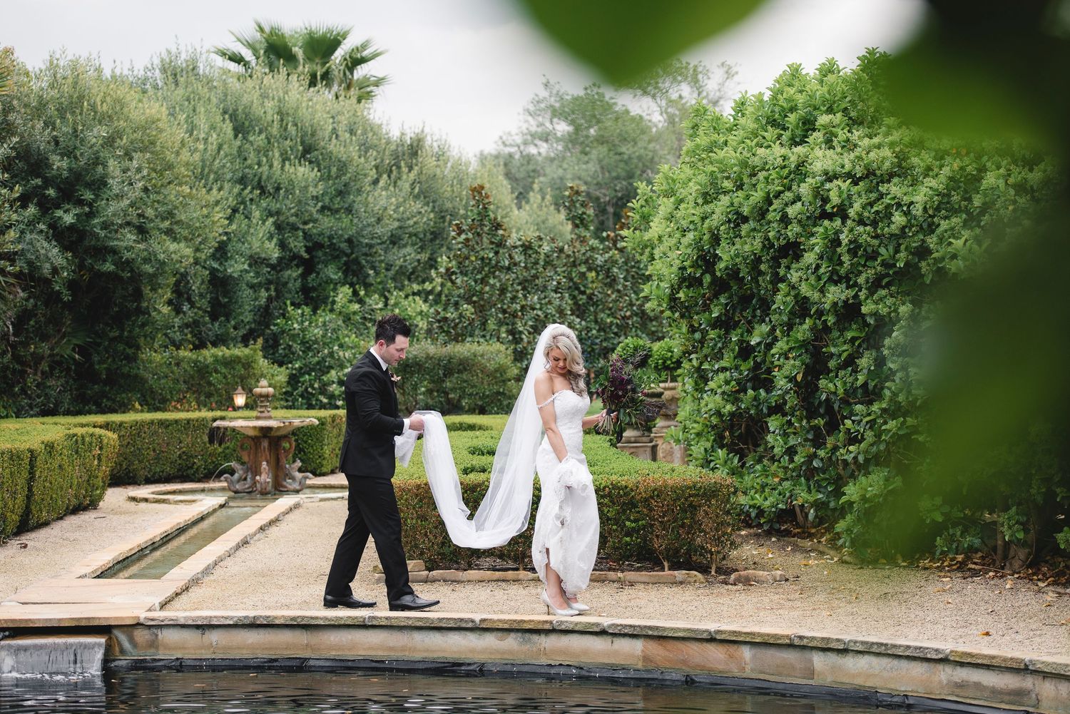 Wedding couple walking along a garden path near a pool surrounded by lush greenery.