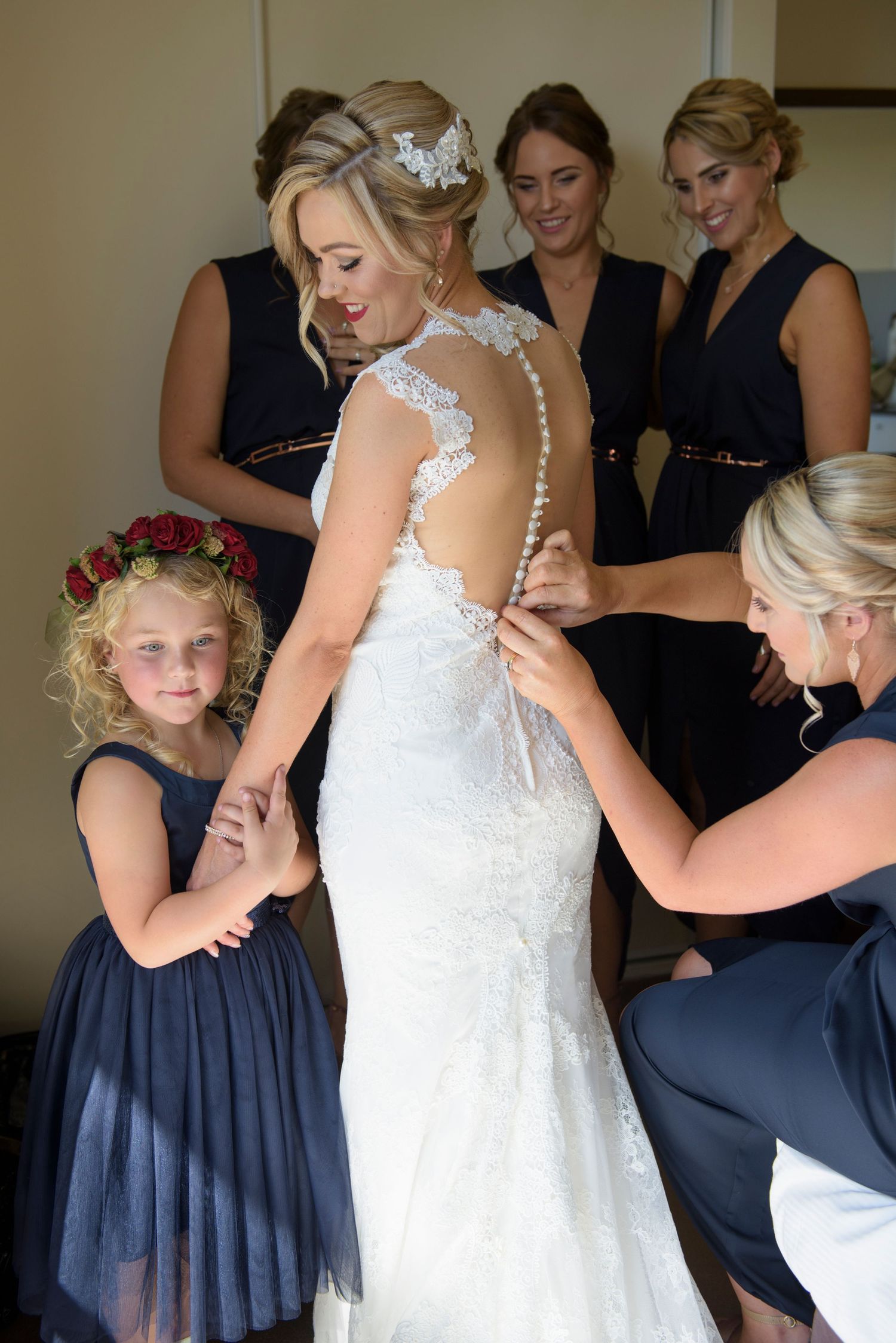 Bridesmaids in navy dresses help the bride get ready by adjusting her white lace wedding gown.