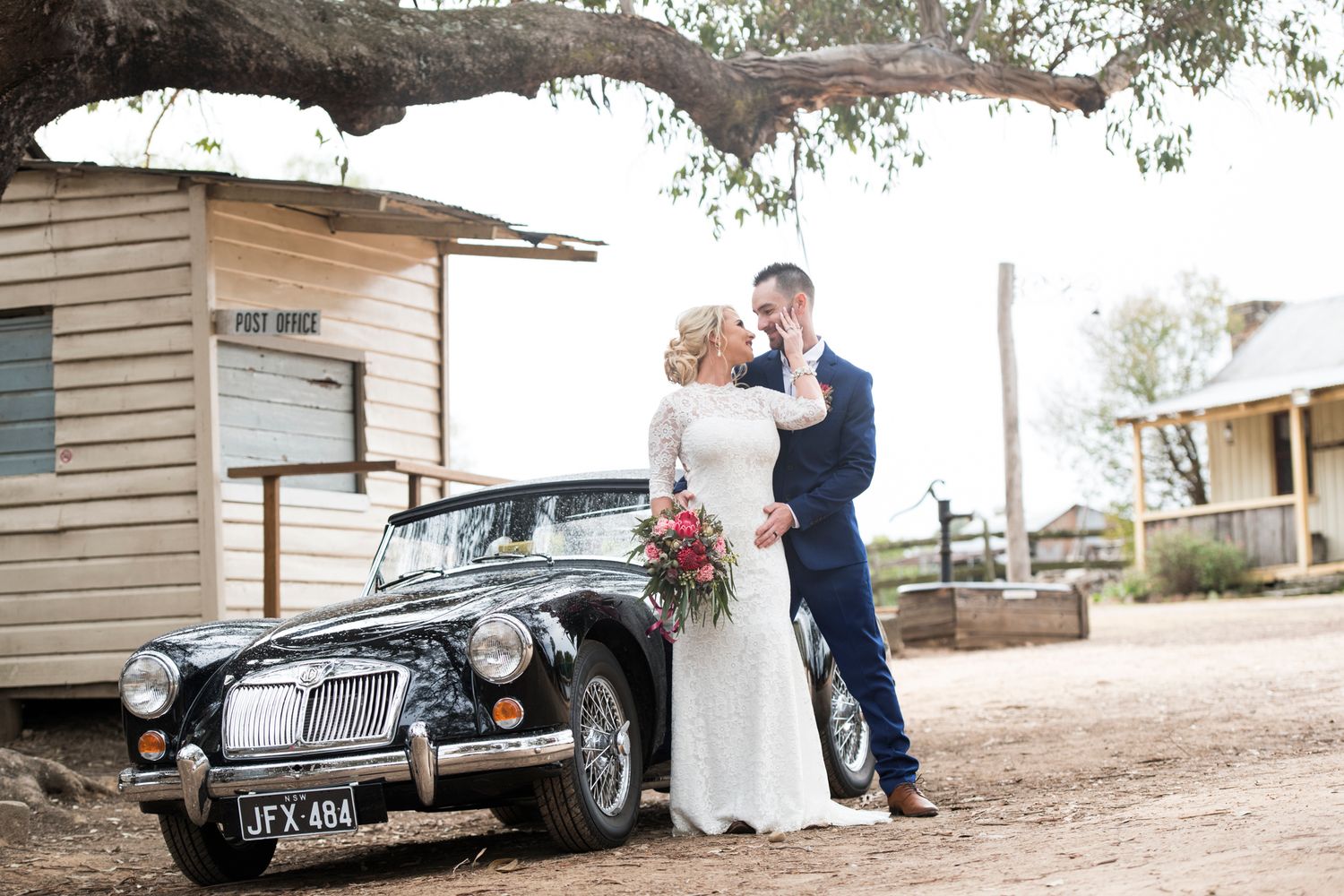 Vintage black car parked outside rustic building with bride and groom posing together.