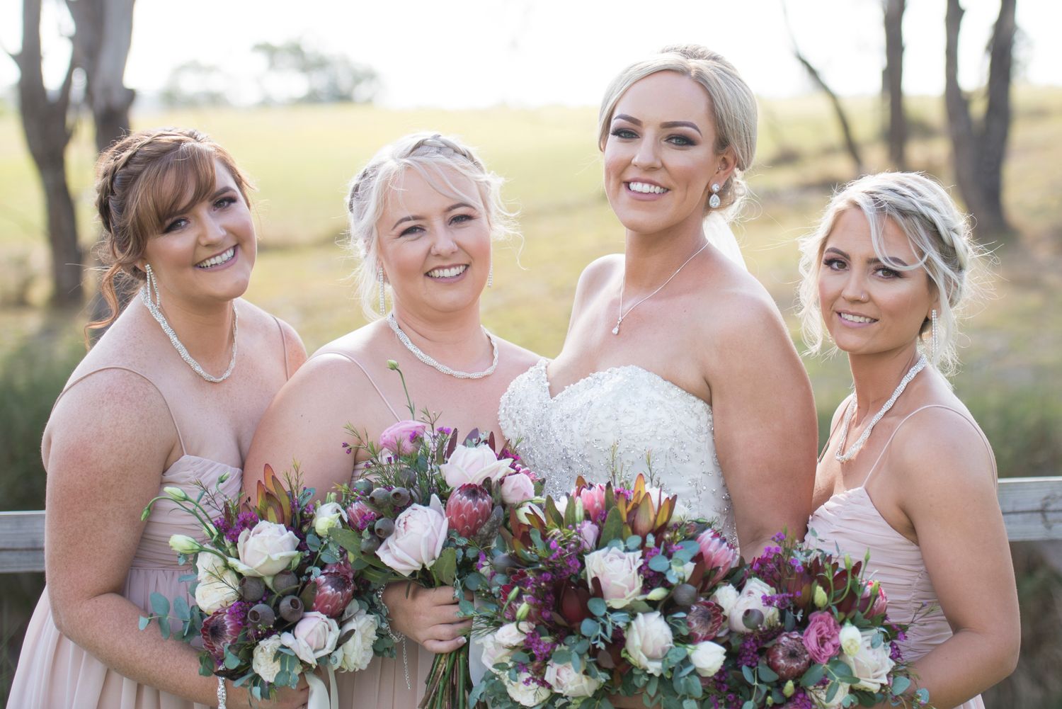 Bride and bridesmaids in blush pink dresses hold pink and white bouquets while smiling outdoors.