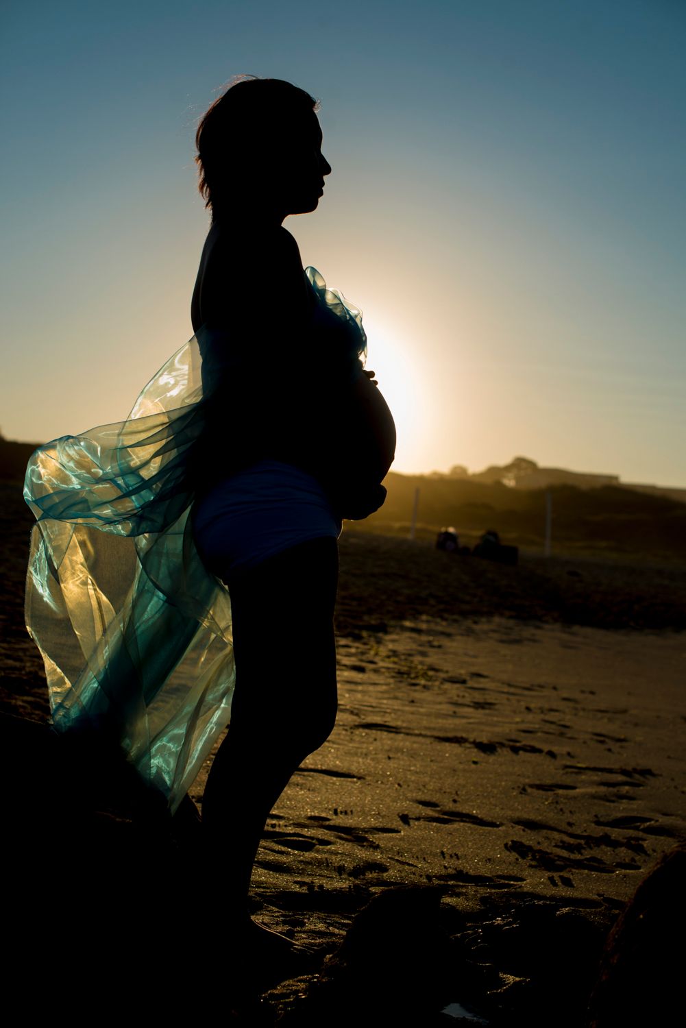 Silhouette figure of mother to be with flowing fabric against sunset on beach with mountains in background.