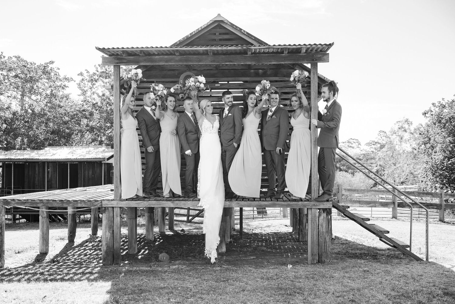 A black and white photo shows a wedding party posing together on a rustic wooden gazebo structure.