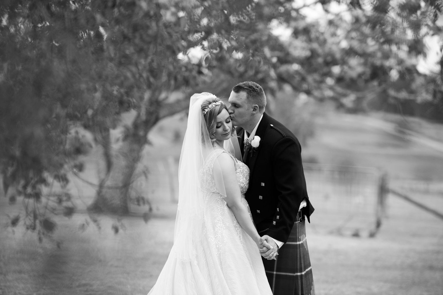 Black and white wedding photo of bride and groom sharing a kiss outdoors, groom wearing traditional Scottish kilt.