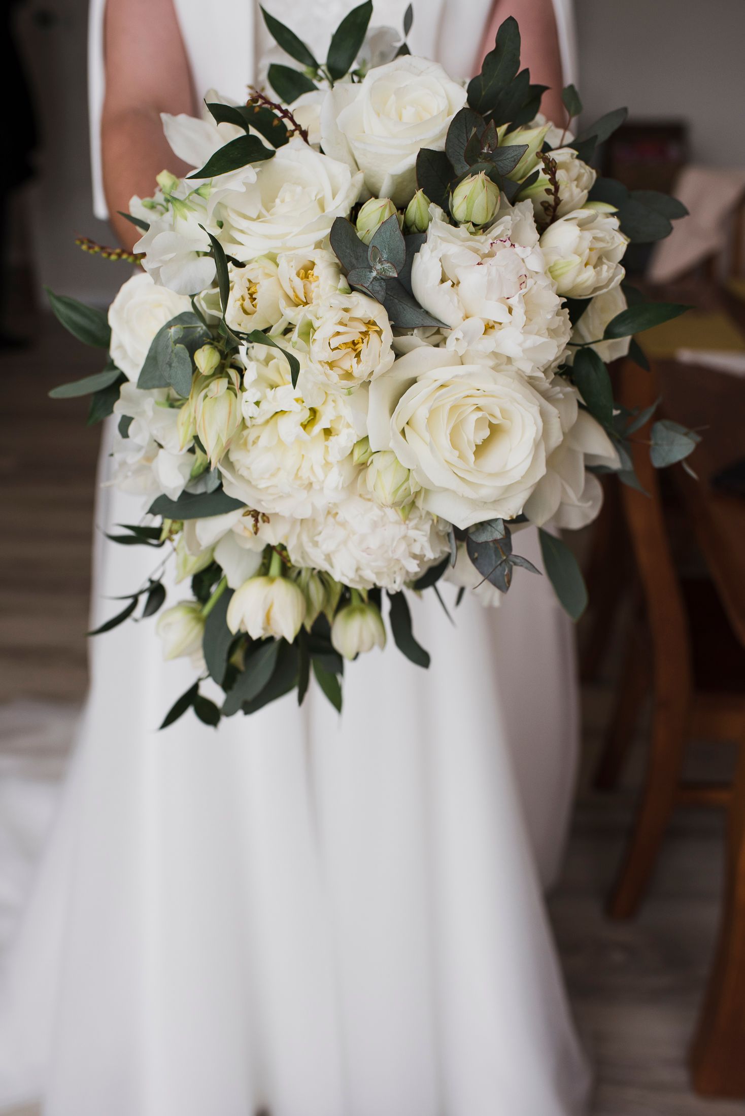 A stunning white bridal bouquet featuring roses, peonies and greenery against a flowing white wedding dress.
