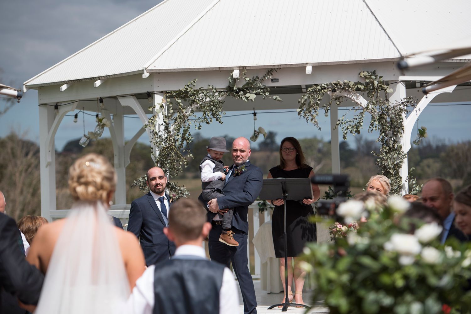 Bride walking down isle. Groom holds son before the wedding ceremony under a white gazebo.