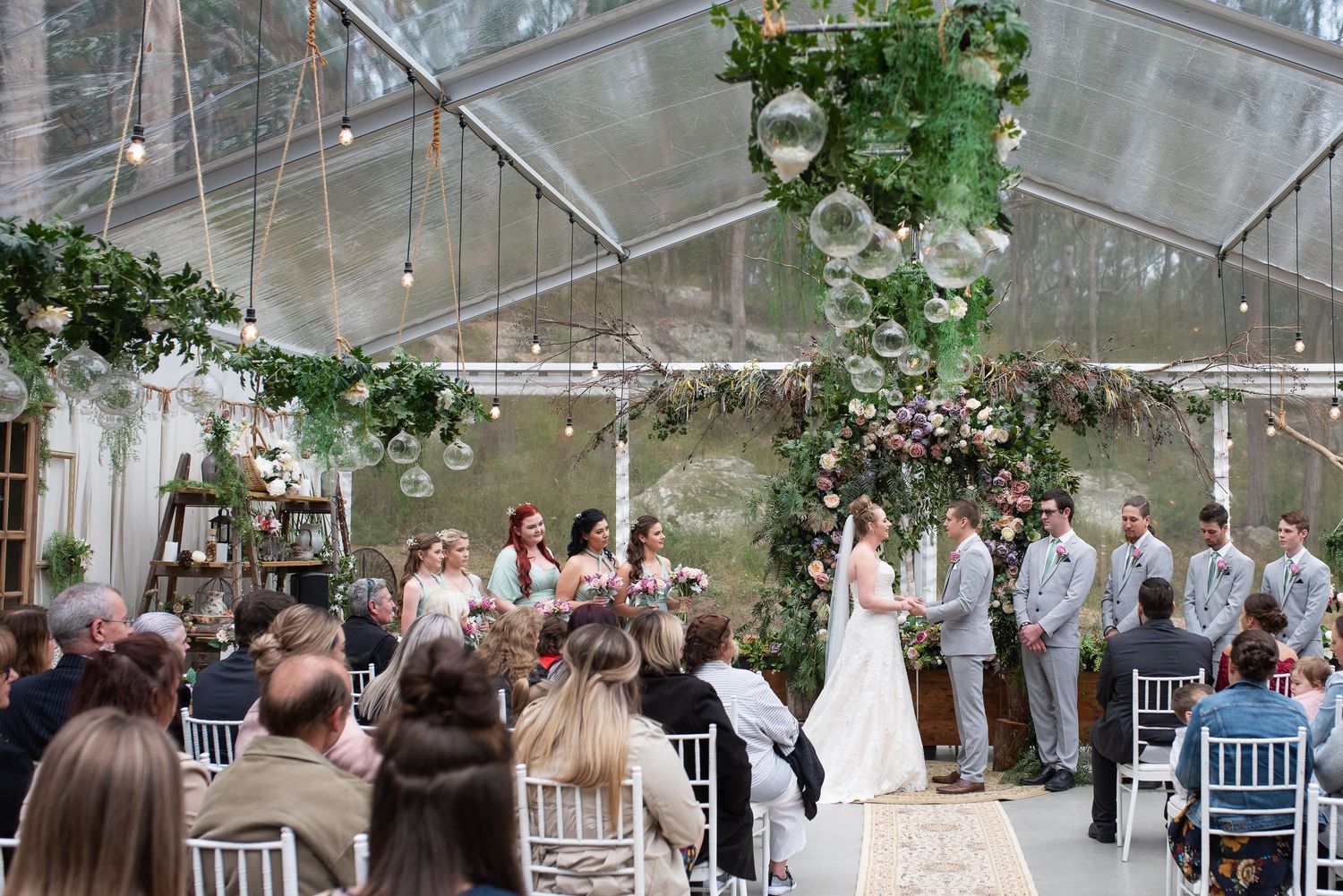 Wedding ceremony taking place, decorated with hanging greenery and string lights.