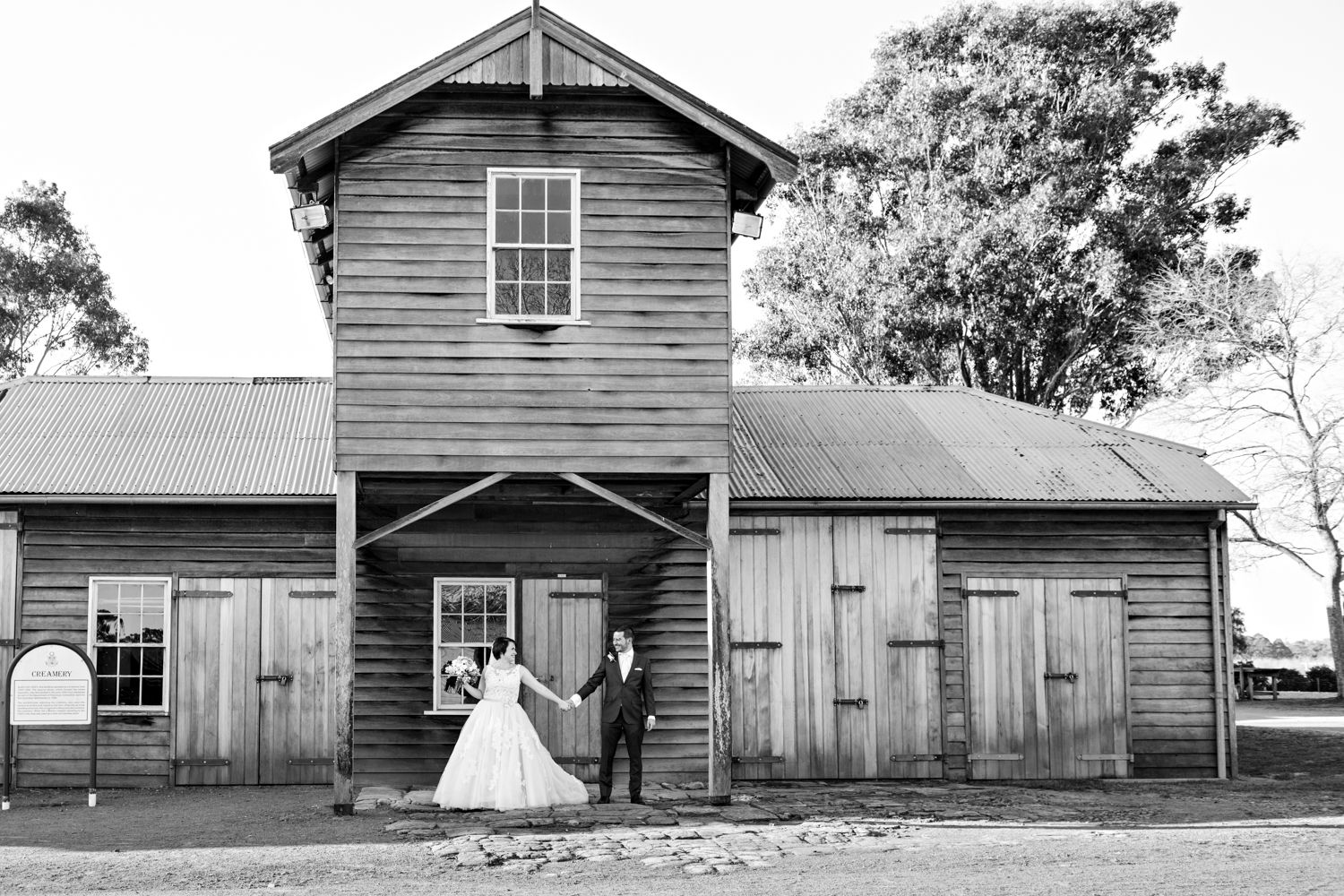 Historic wooden barns and outbuildings stand in a row at a heritage farm site with bride and groom.