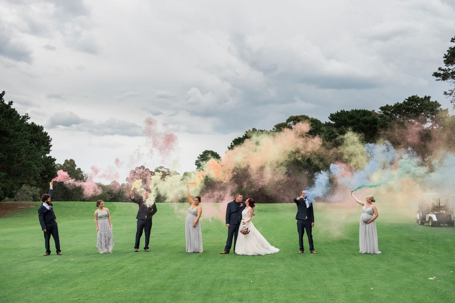 Wedding party poses with colorful smoke bombs in an outdoor field setting.