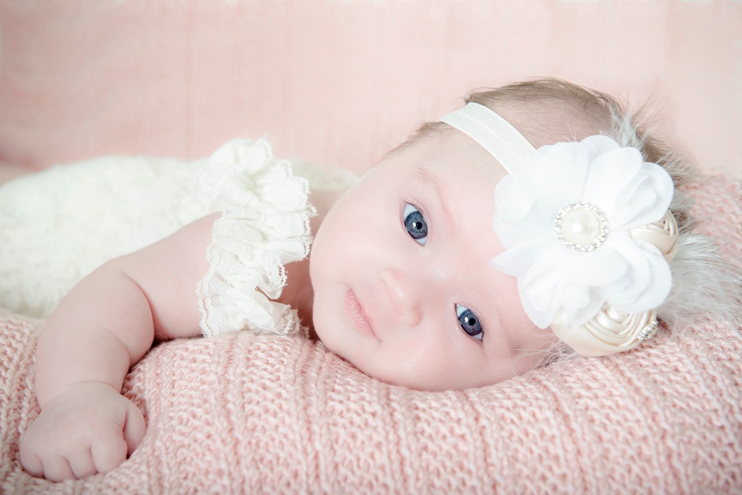 Baby wearing white floral headband resting on pink knit blanket.