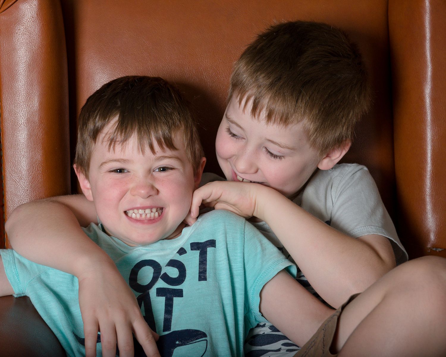 Children laughing and hugging on brown leather couch during candid moment.