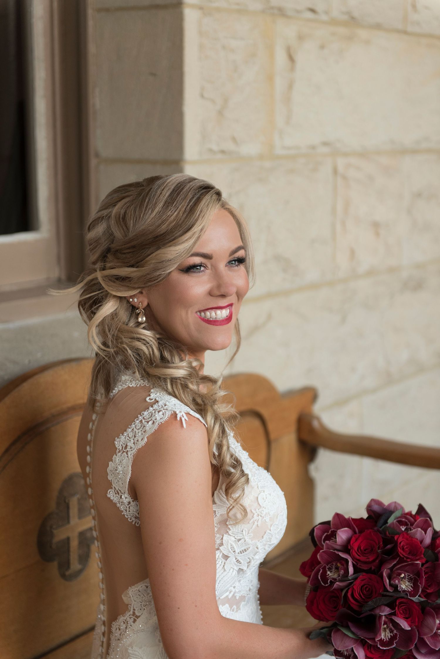 Bride in white lace dress holding deep red roses against stone wall.