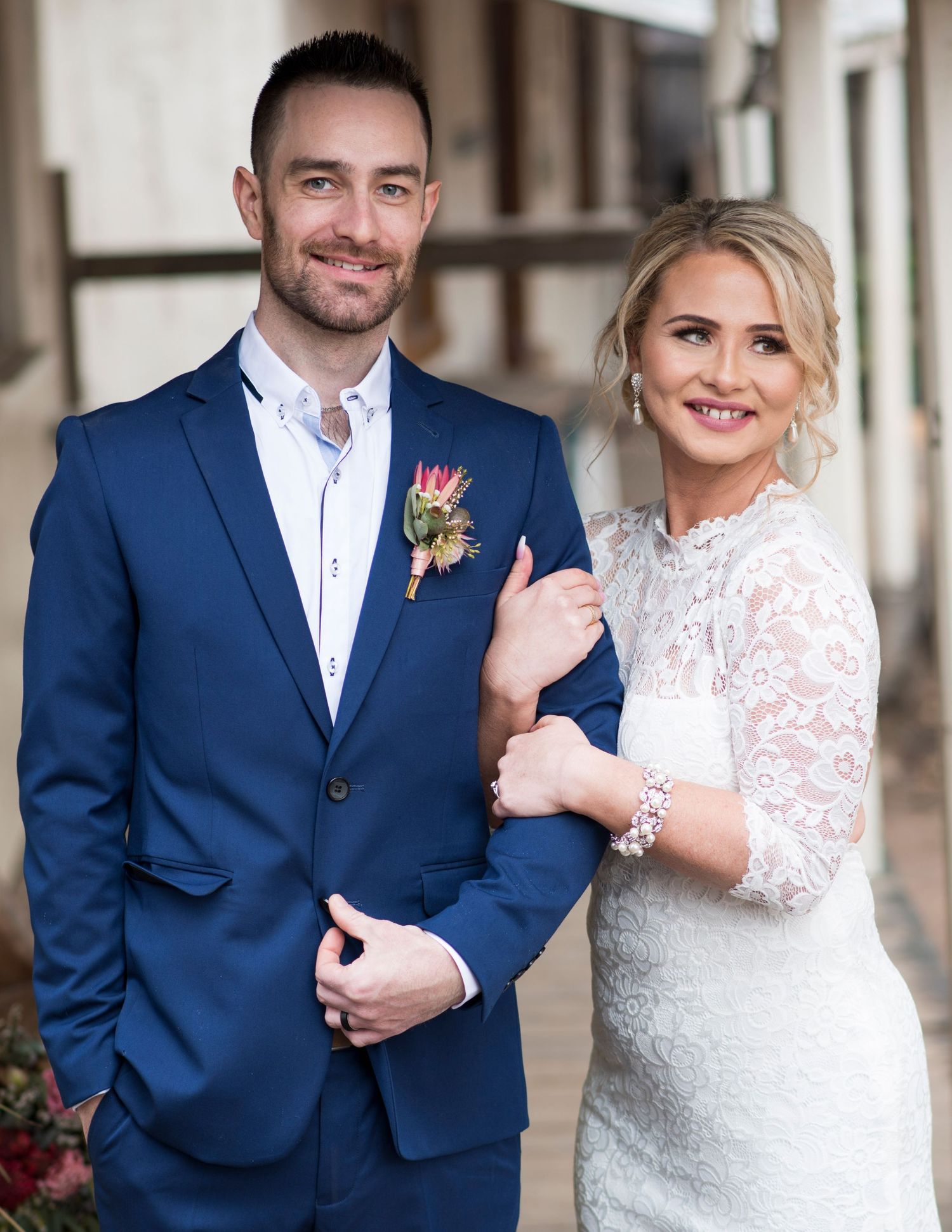 Wedding couple poses in elegant attire against rustic architectural backdrop.
