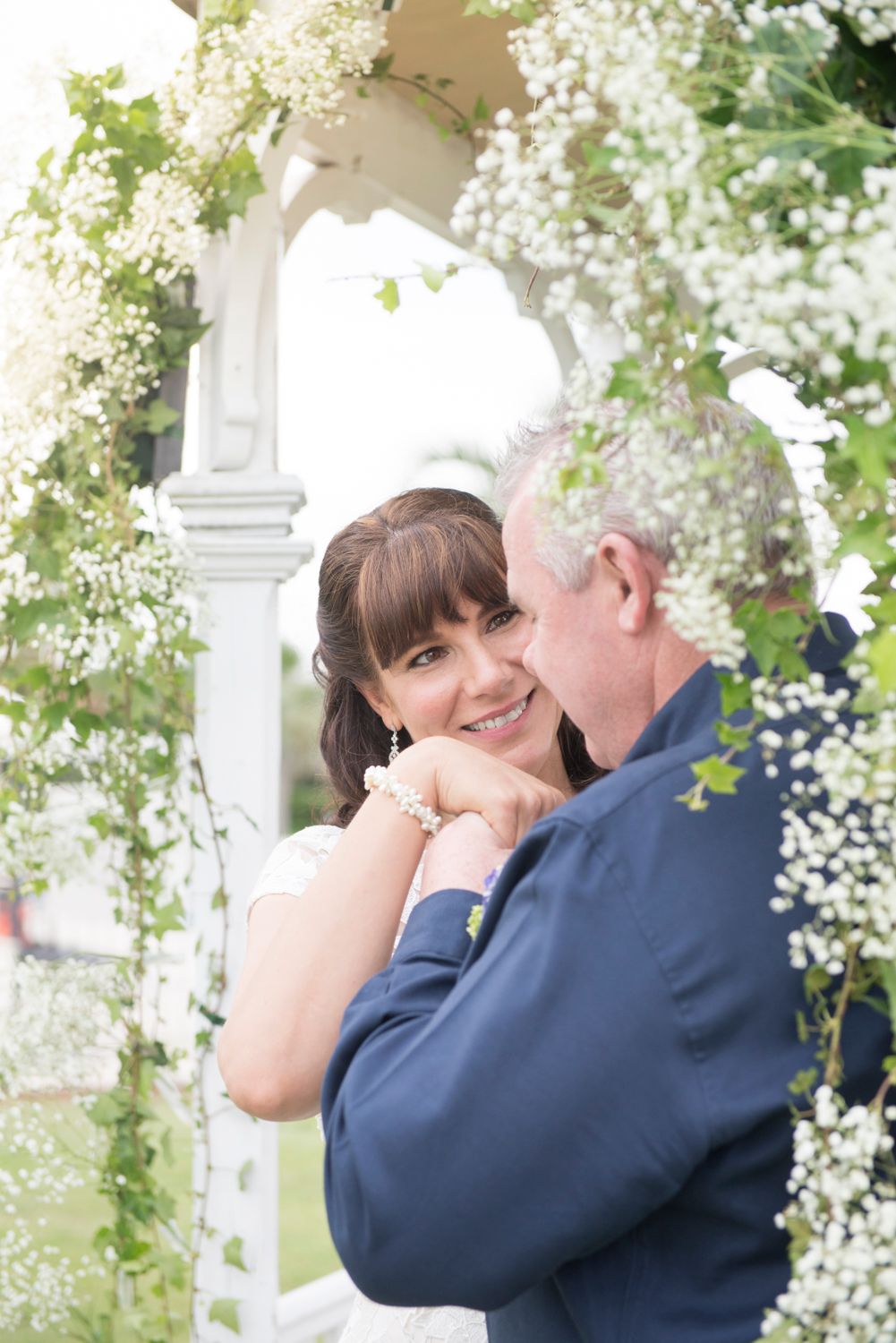 Romantic wedding couple embraces under a white floral arbor in an outdoor garden setting.