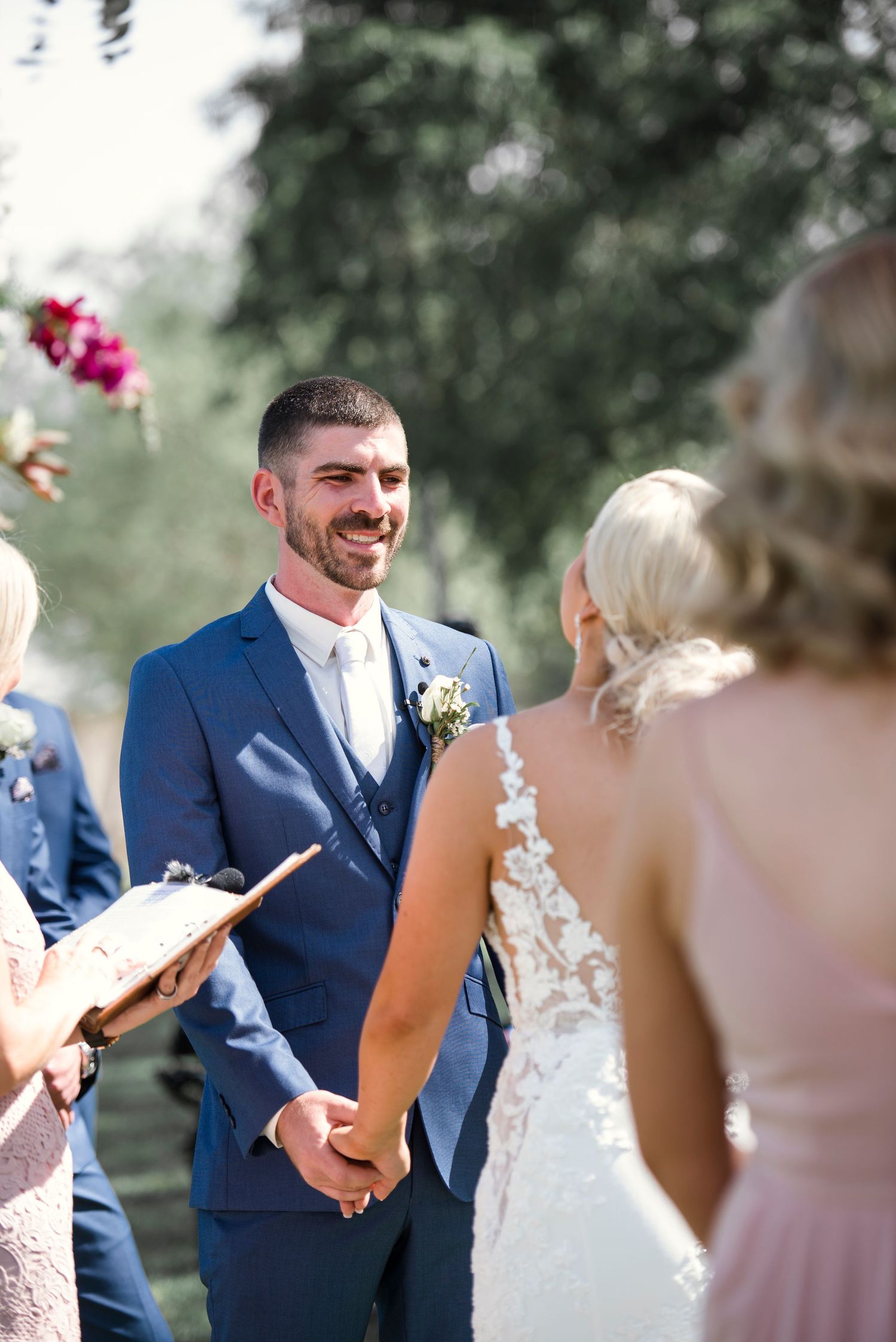 Groom in navy suit smiling at bride while holding hands during outdoor wedding ceremony.
