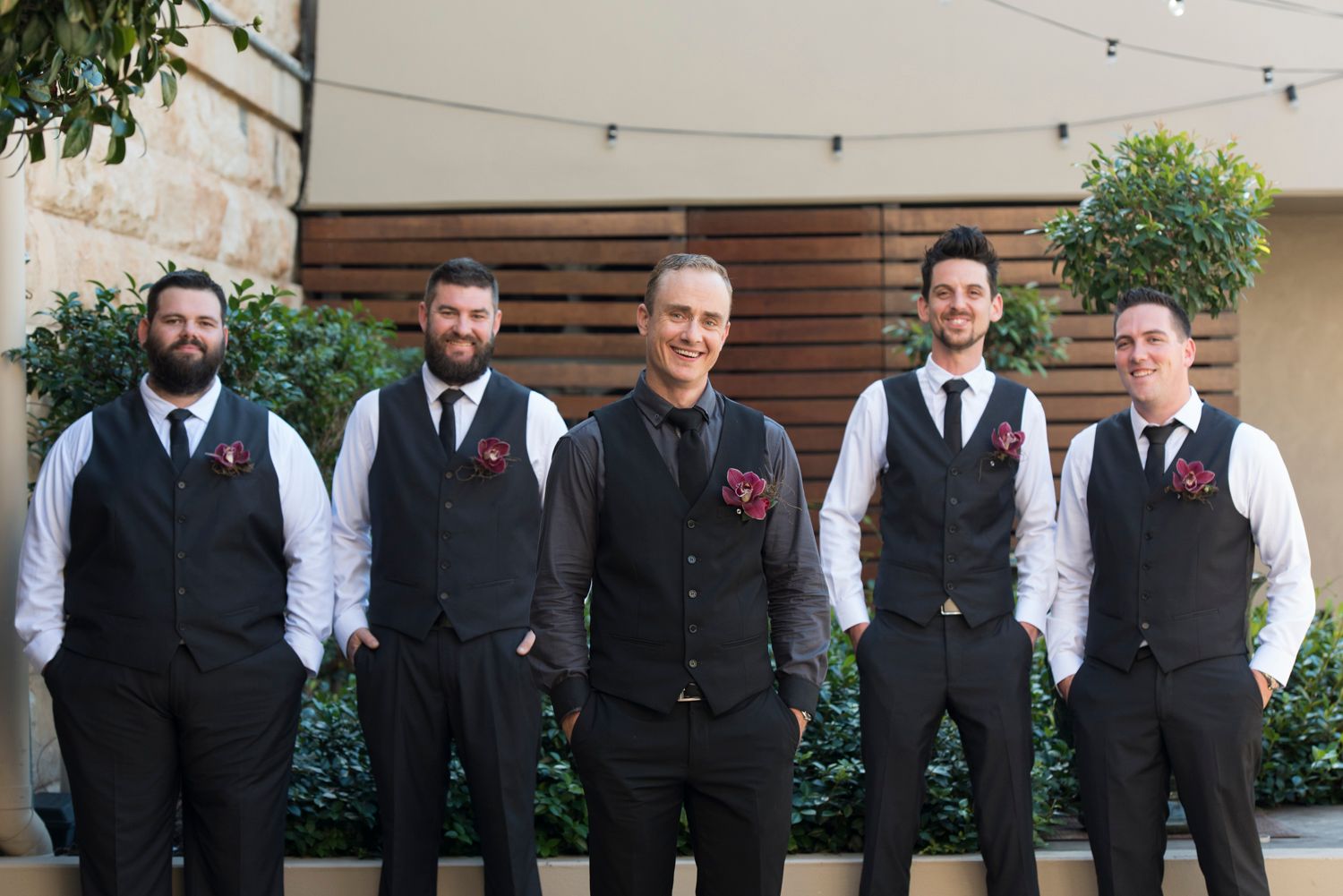 Groomsmen in matching black vests and white shirts stand in a line outdoors.