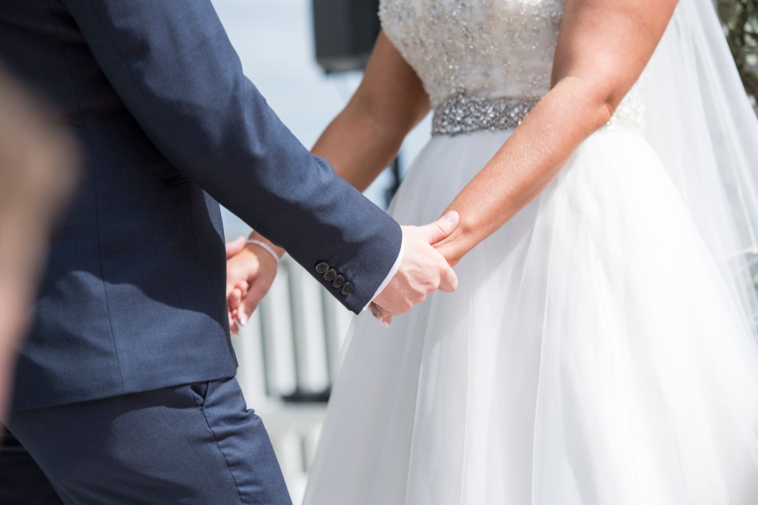A couple holds hands during their ceremony, with the bride in white and groom in navy blue.