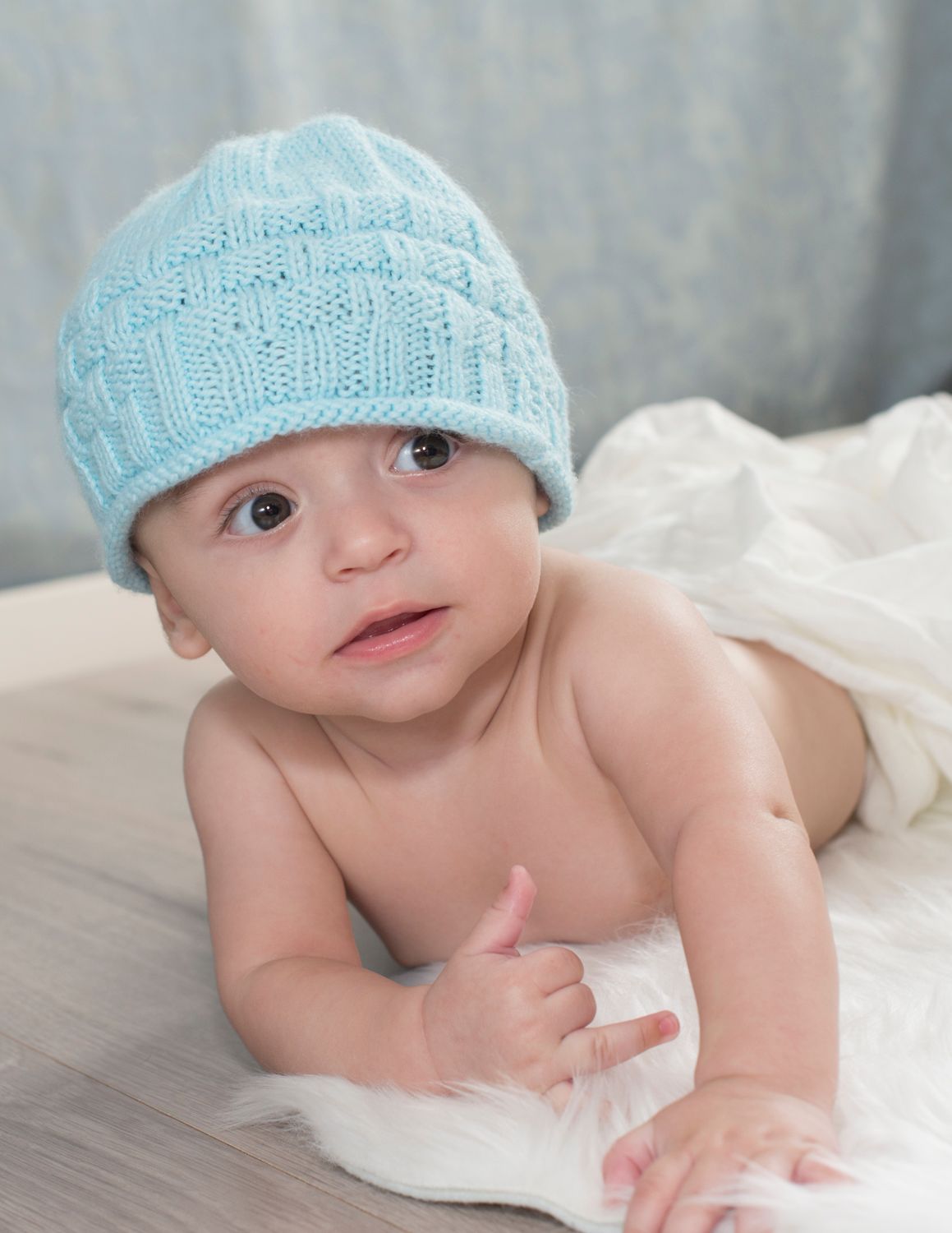 Baby wearing light blue knit hat lies on white fabric while looking at camera.