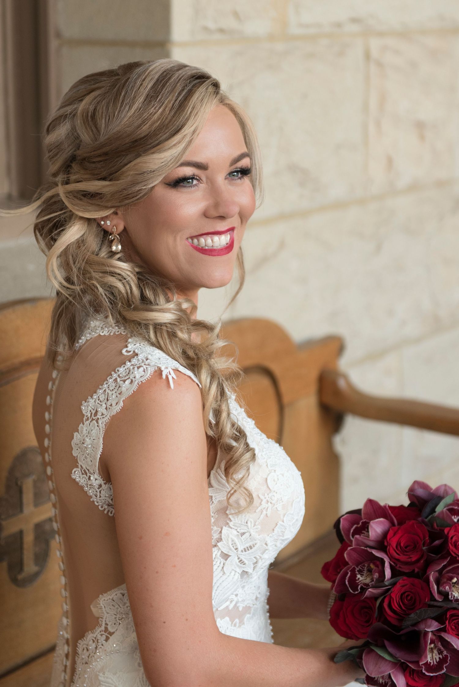Bride in white lace dress holding deep red roses against stone wall.