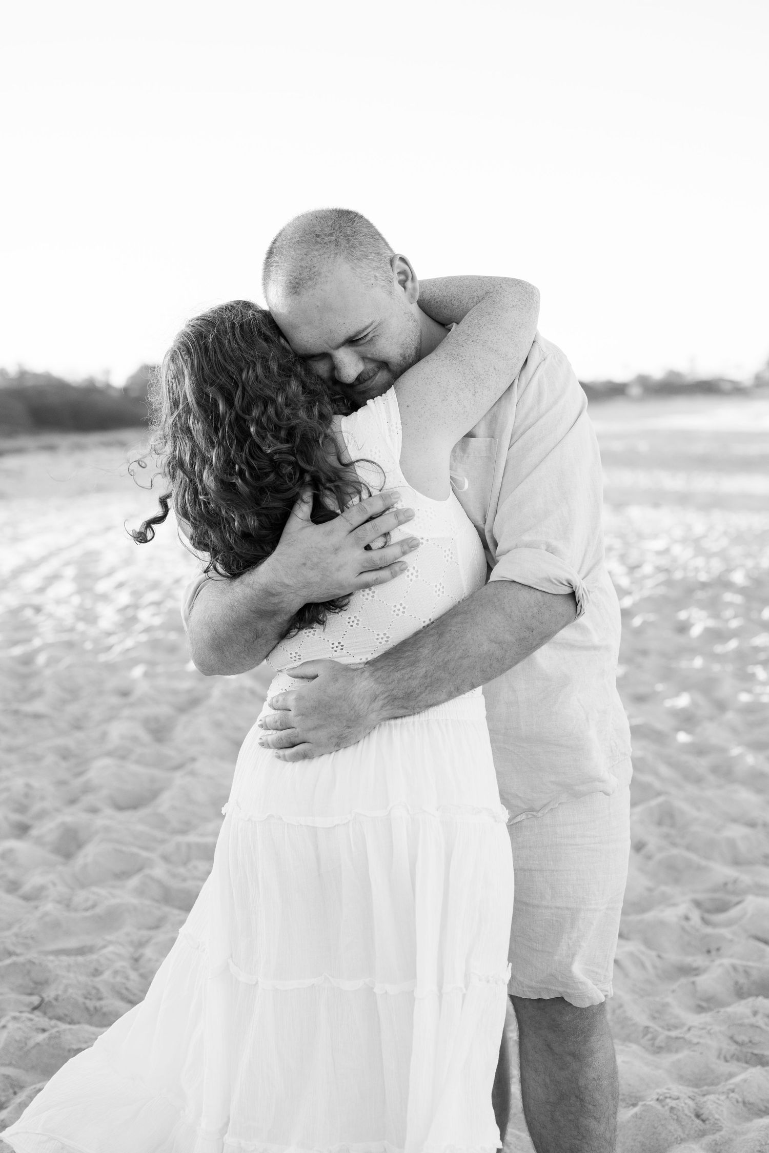 Black and white photo of a couple embracing tenderly on a sandy beach, woman in white dress, man in light shirt.