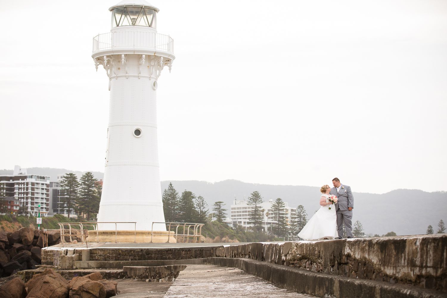 A couple stands on a stone path near the white Wollongong Head Lighthouse with mountains in the background.