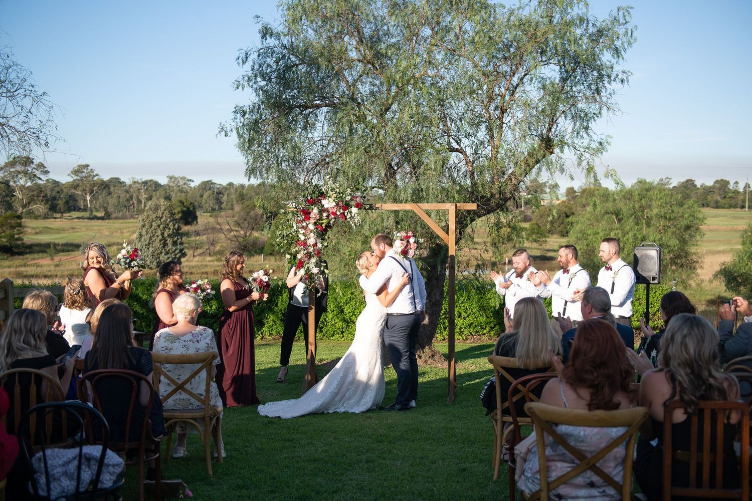 Wedding ceremony guests gather outdoors under eucalyptus trees in a rustic countryside setting during golden hour.