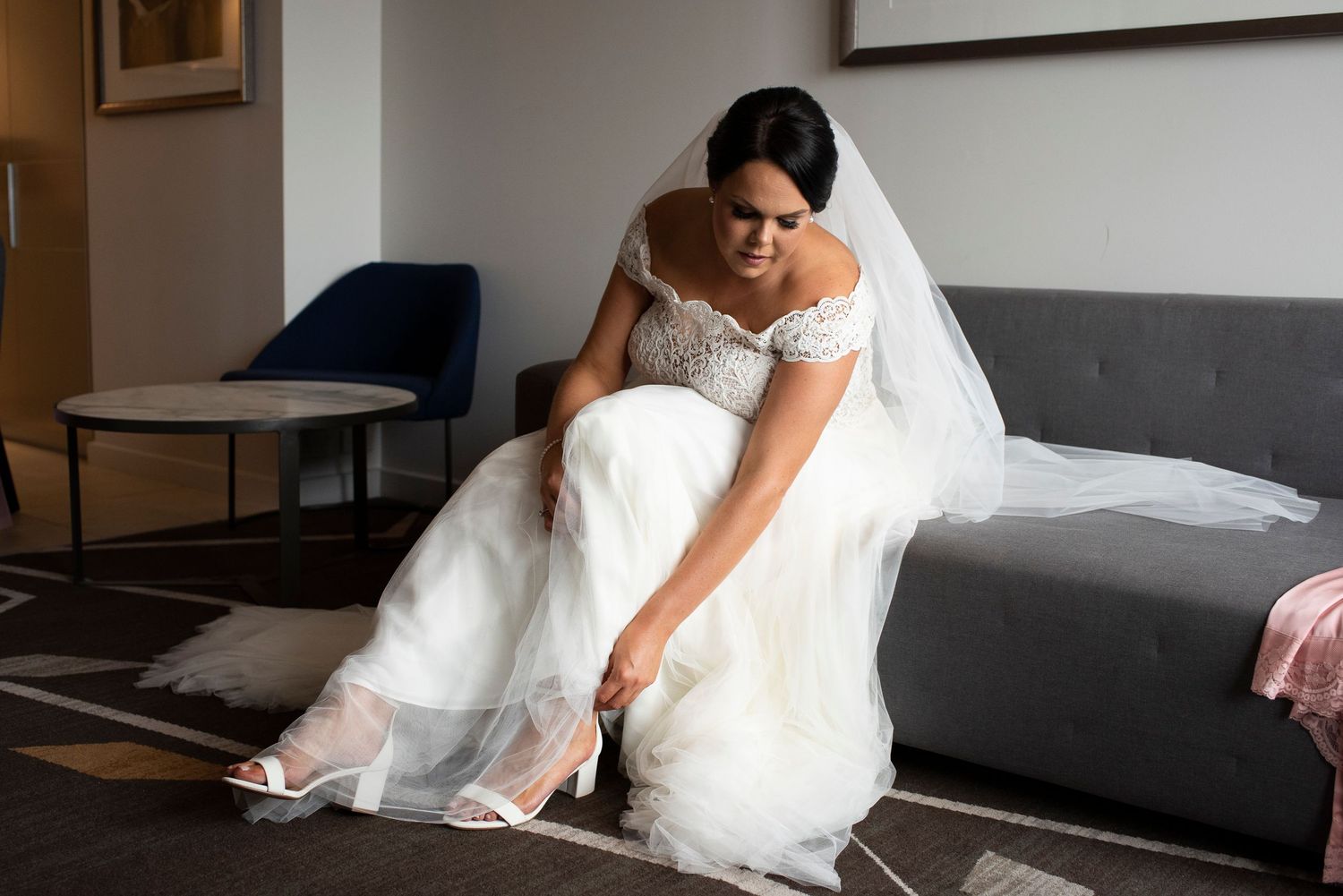 Bride in white dress sits on modern grey couch during indoor photoshoot.