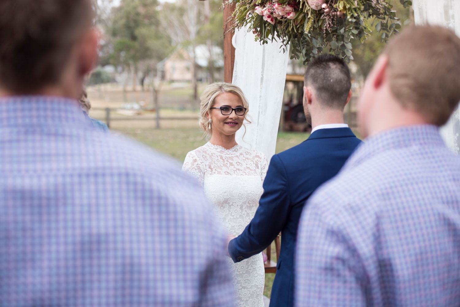 A wedding ceremony takes place outdoors with guests watching as a couple exchanges vows under a native arch.
