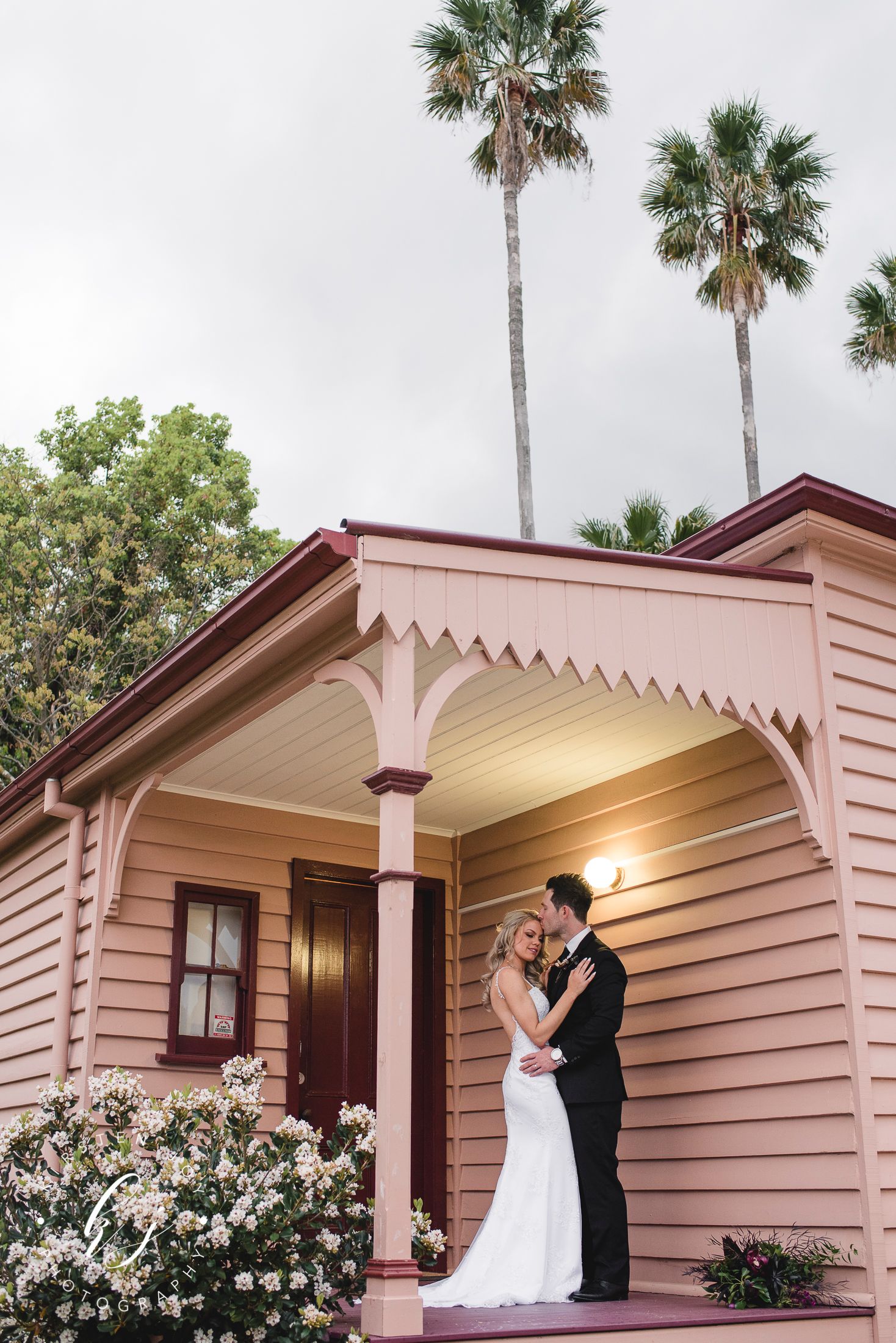 A couple shares an intimate moment on the porch of a pink Victorian-style house with palm trees.