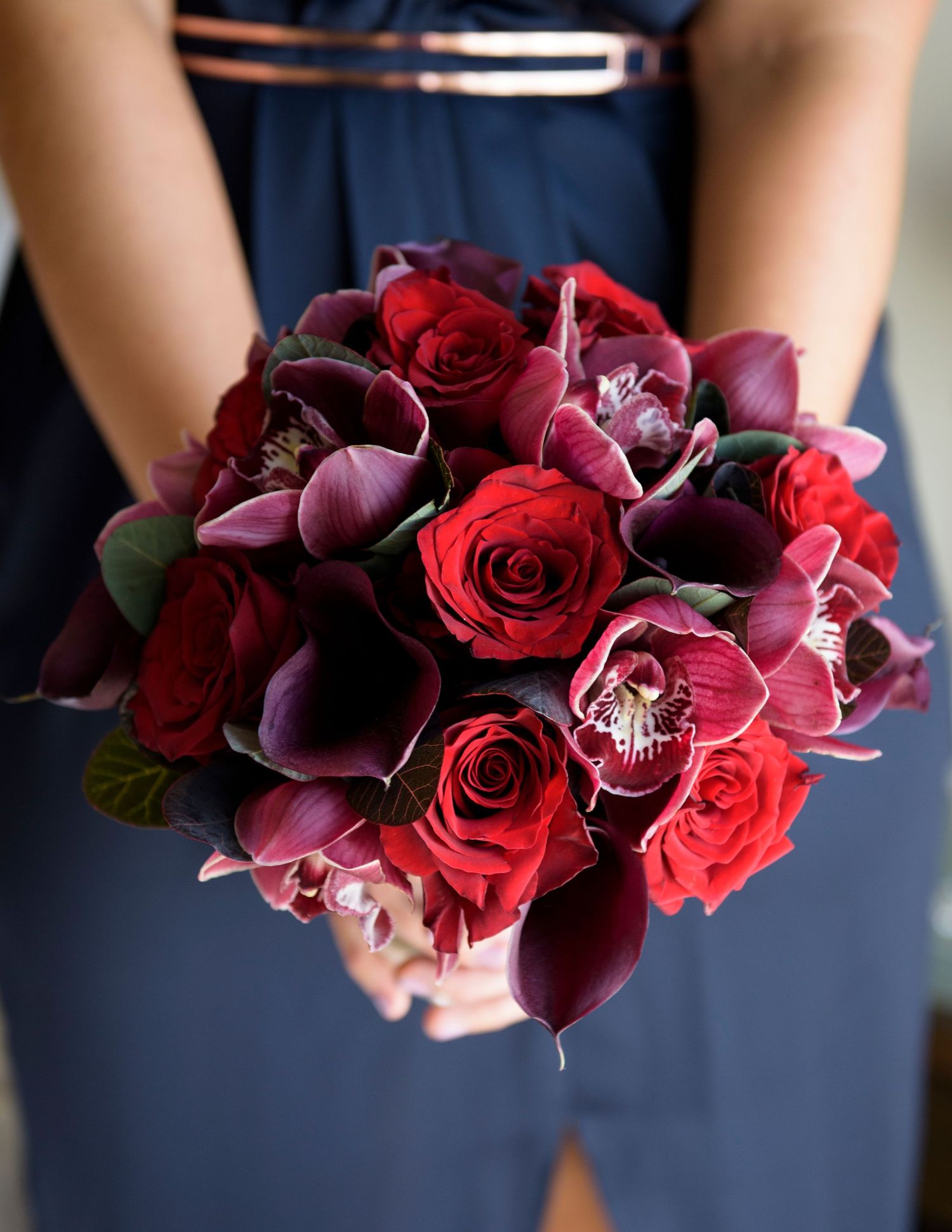 A bridesmaid holds a stunning bouquet of deep red and burgundy roses with dark purple accents against a navy dress.